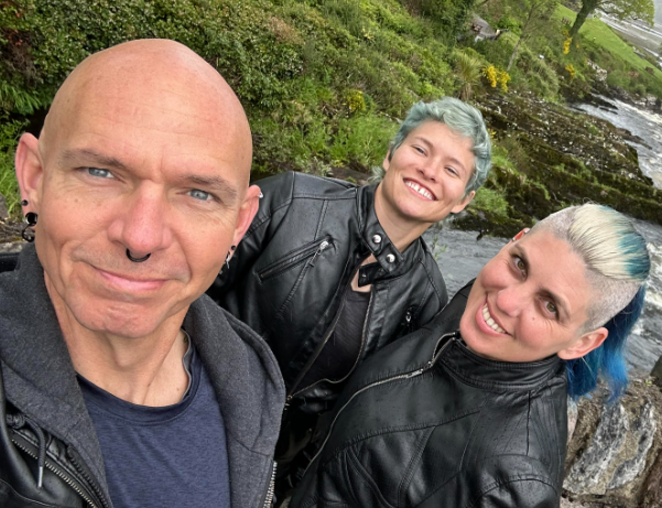 Color selfie of three adults standing close together outdoors on a stone bridge overlooking a fast-moving river and lush green landscape. On the left, a male presenting person with a shaved head and facial piercings looks at the camera. In the center, a female presenting person with short, light teal hair smiles broadly, wearing a black leather jacket. On the right, a female presenting person with shaved sides and longer hair dyed blue and blonde smiles toward the camera, also wearing a black leather jacket. The river flows over rocks behind them, with trees and rolling hills in the background.