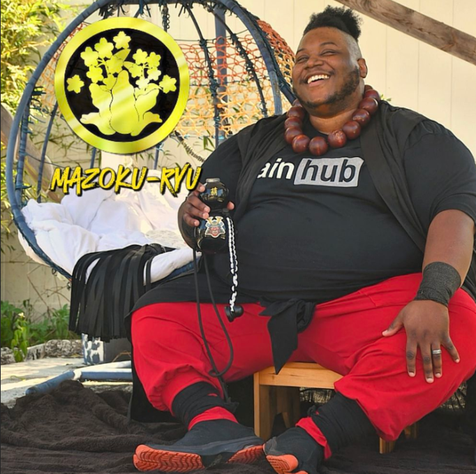 A smiling person sits outdoors on a low wooden stool in front of a hanging egg chair, wearing a black “painhub” T-shirt, red pants, black wrist wraps, and a large beaded necklace. They hold a black bottle with a cord attached. A yellow circular logo with a stylized tree and the text “Mazoku-ryu” appears in the upper left corner.