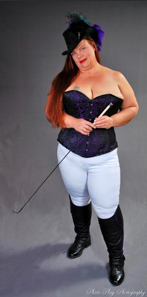 Studio portrait of a female presenting person standing against a plain gray backdrop. They wear a black top hat decorated with feathers, a purple corset, light-colored pants, and tall black boots. Their long red hair falls over one shoulder. They hold a riding crop in both hands while looking toward the camera with a confident expression.