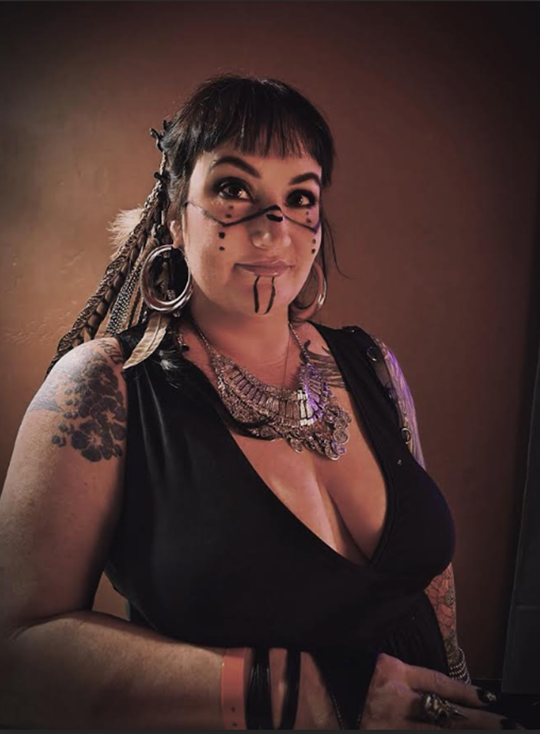 Portrait of a female presenting person standing indoors against a warm-toned wall. They have short dark hair with blunt bangs and decorative braids with feathers. Their face features stylized black face paint with dots and lines across the cheeks, nose, and chin. They wear large hoop earrings, layered metallic necklaces, and a sleeveless black top. Tattoos are visible on their arms and shoulder as they look directly at the camera with a calm expression.