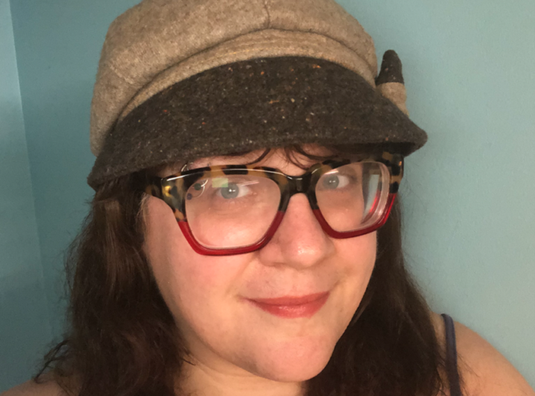 Close-up portrait of a female presenting person standing against a teal wall. They have long dark hair and wear large tortoiseshell-style glasses with red lower frames and a gray cap with a short brim. They look slightly off to the side with a soft, neutral expression.
