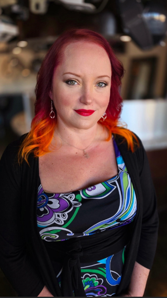 Color portrait of a female presenting person looking directly at the camera. They have shoulder-length hair dyed red with bright orange ends and wear red lipstick. They are dressed in a patterned dress with blue, green, purple, and black designs and a black cardigan. The background appears to be an indoor setting with softly blurred lights and equipment.