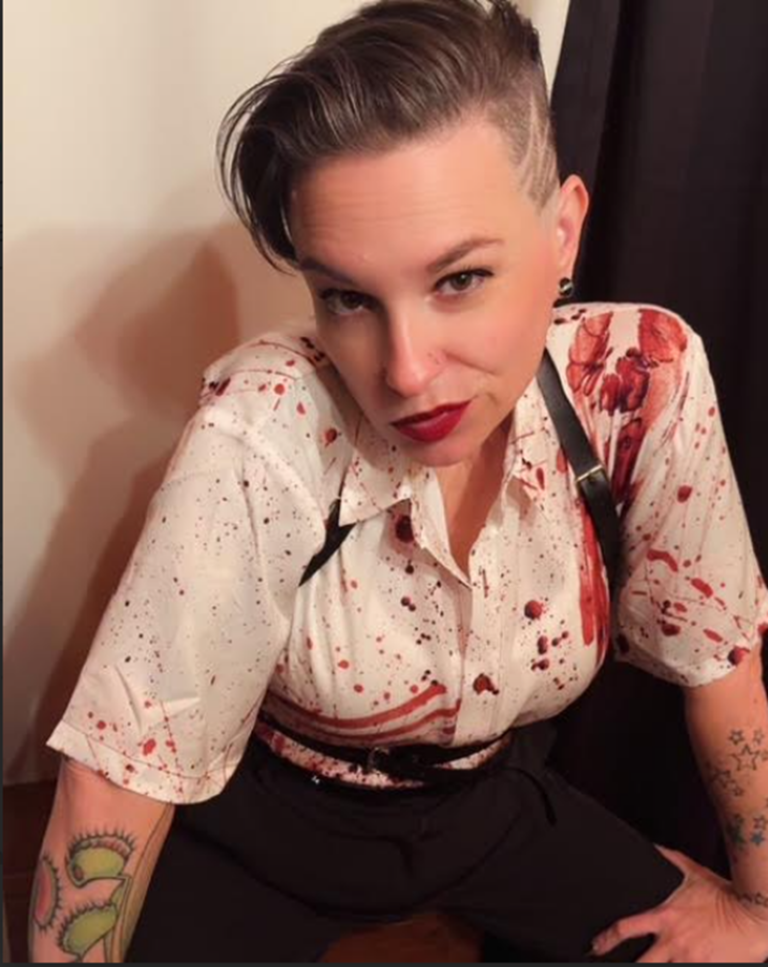 Portrait of a nonbinary presenting person leaning slightly forward while looking directly at the camera. They have short hair styled with shaved sides and longer hair swept to one side. They wear a white button-up shirt patterned with red splatter effects, dark suspenders, and dark pants. Visible tattoos appear on their forearms. The background shows a neutral indoor wall and a dark curtain.