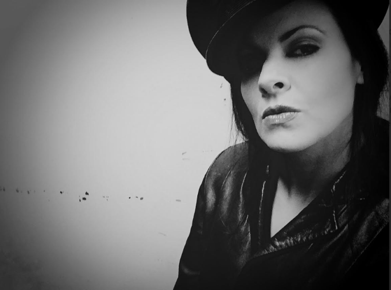 Black-and-white portrait of a person wearing a dark hat and a leather jacket. They face the camera at a slight angle with a composed expression, dark hair falling to one side. The background is a plain wall, creating a dramatic, high-contrast look.