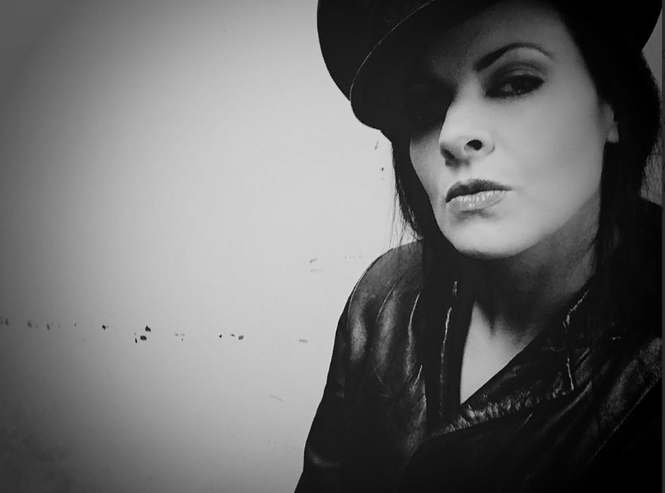 Black-and-white portrait of a person wearing a dark hat and a leather jacket. They face the camera at a slight angle with a composed expression, dark hair falling to one side. The background is a plain wall, creating a dramatic, high-contrast look.