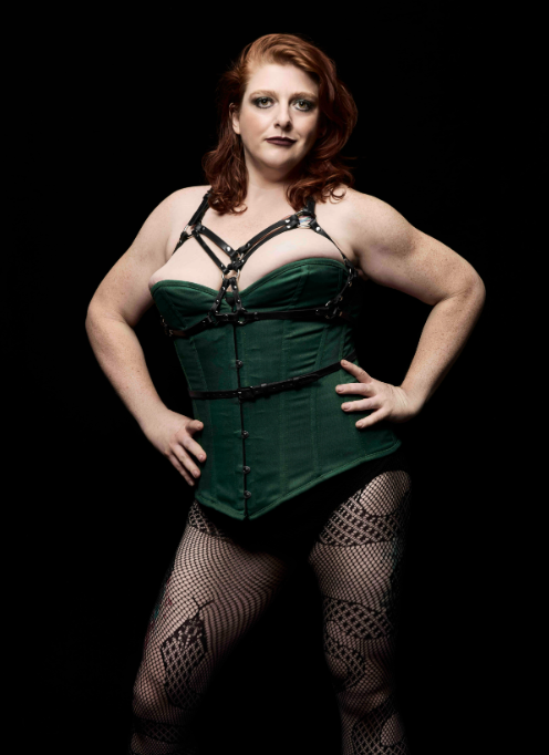 Full-body studio portrait of a red-haired woman posing with her hands on her hips. She wears a dark green corset with a black harness, black briefs, and patterned fishnet tights against a black background.