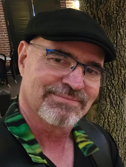 Outdoor close-up of a smiling man wearing glasses and a black flat cap. He has a gray goatee and wears a black shirt with a green patterned collar.