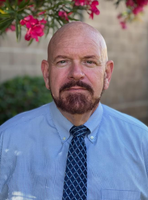 Outdoor headshot of a bald man with a trimmed brown beard, wearing a light blue dress shirt and patterned navy tie. Pink flowers and greenery appear softly blurred behind him.