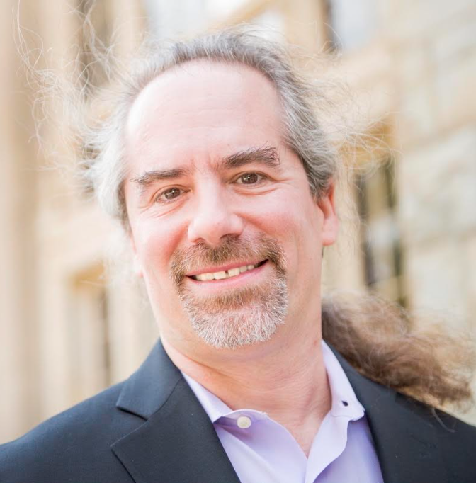 Outdoor headshot of Dr. Brad Sagarin smiling at the camera. He has long gray hair tied back, a gray goatee, and wears a dark blazer over a light purple shirt.