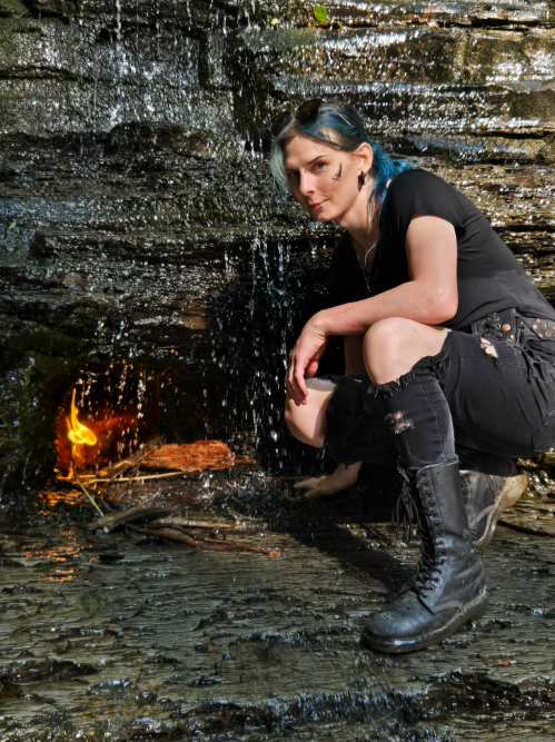 A person with short blue hair crouches beside a small eternal flame beneath a rocky waterfall, wearing torn black clothing and knee-high lace-up boots.