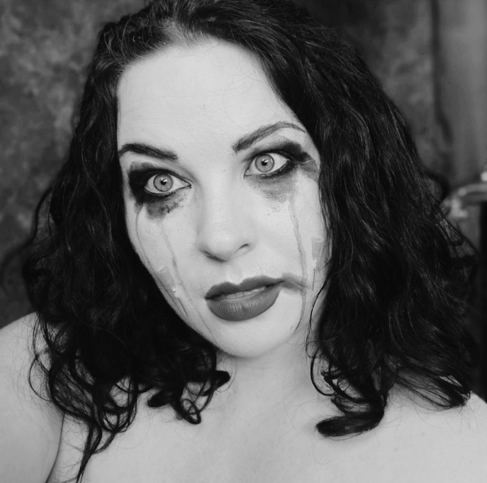 Black-and-white close-up portrait of a person with shoulder-length dark hair, dramatic eye makeup, and dark streaks resembling tears running down her cheeks. They gaze off to the side.