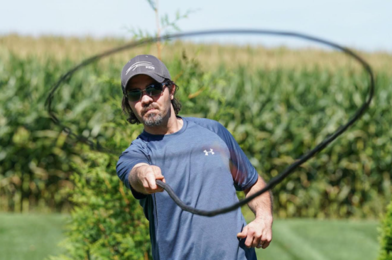 A man wearing sunglasses, a baseball cap, and a blue athletic shirt swings a long black whip outdoors beside a cornfield.