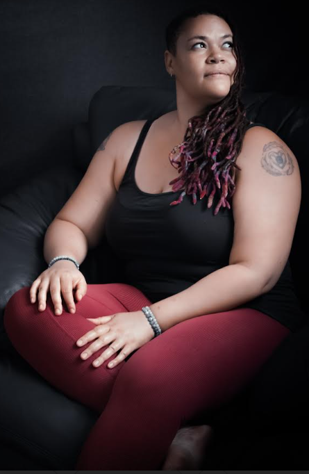 Portrait of a seated Black woman wearing a black tank top and burgundy pants. She has long locs with pink accents, a tattoo on her upper arm, and looks thoughtfully toward the light.
