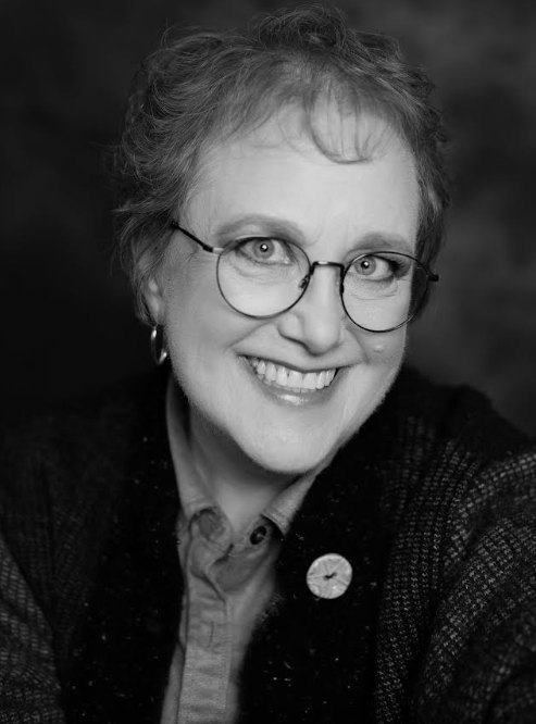 Black-and-white portrait of a smiling person with short, wavy hair and round glasses, wearing hoop earrings, a collared shirt, and a dark textured jacket with a circular pin.