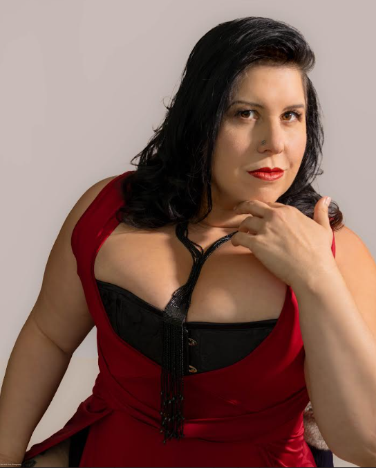 Studio portrait of a woman with shoulder-length black hair and red lipstick, wearing a low-cut red dress over a black corset. She rests one hand beneath her chin and gazes to the side.