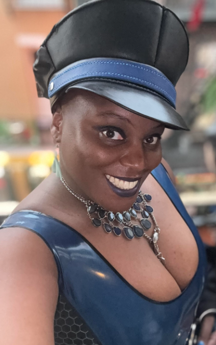 A smiling Black woman wears a shiny blue outfit, a black leather cap with blue trim, and a layered blue statement necklace. She looks directly at the camera.