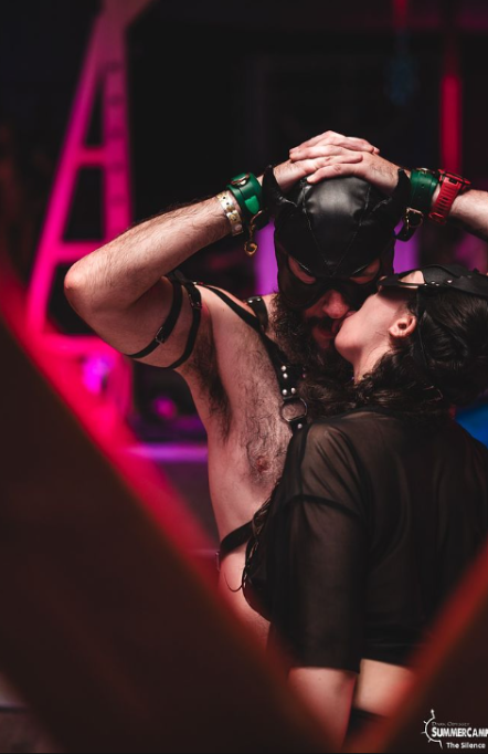Two people in black leather fetish attire share an intimate kiss beneath pink and purple stage lighting. One wears a leather hood and chest harness, while the other wears a sheer top and peaked cap.