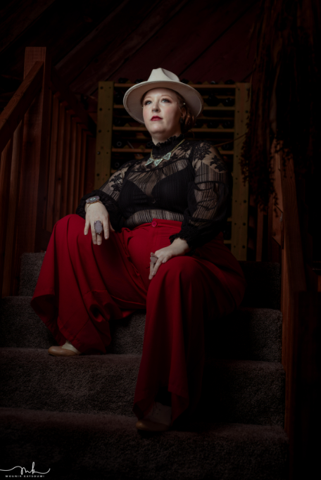 A person sits on a carpeted wooden staircase in a dimly lit room, gazing upward to the left. They wear a cream-colored brimmed hat, a sheer black blouse with floral lace and a high neckline, red wide-leg trousers, and tan shoes. A statement necklace, wristwatch, and rings complete their outfit. Their hands rest on their thighs, with wooden railings and a wine rack behind them.